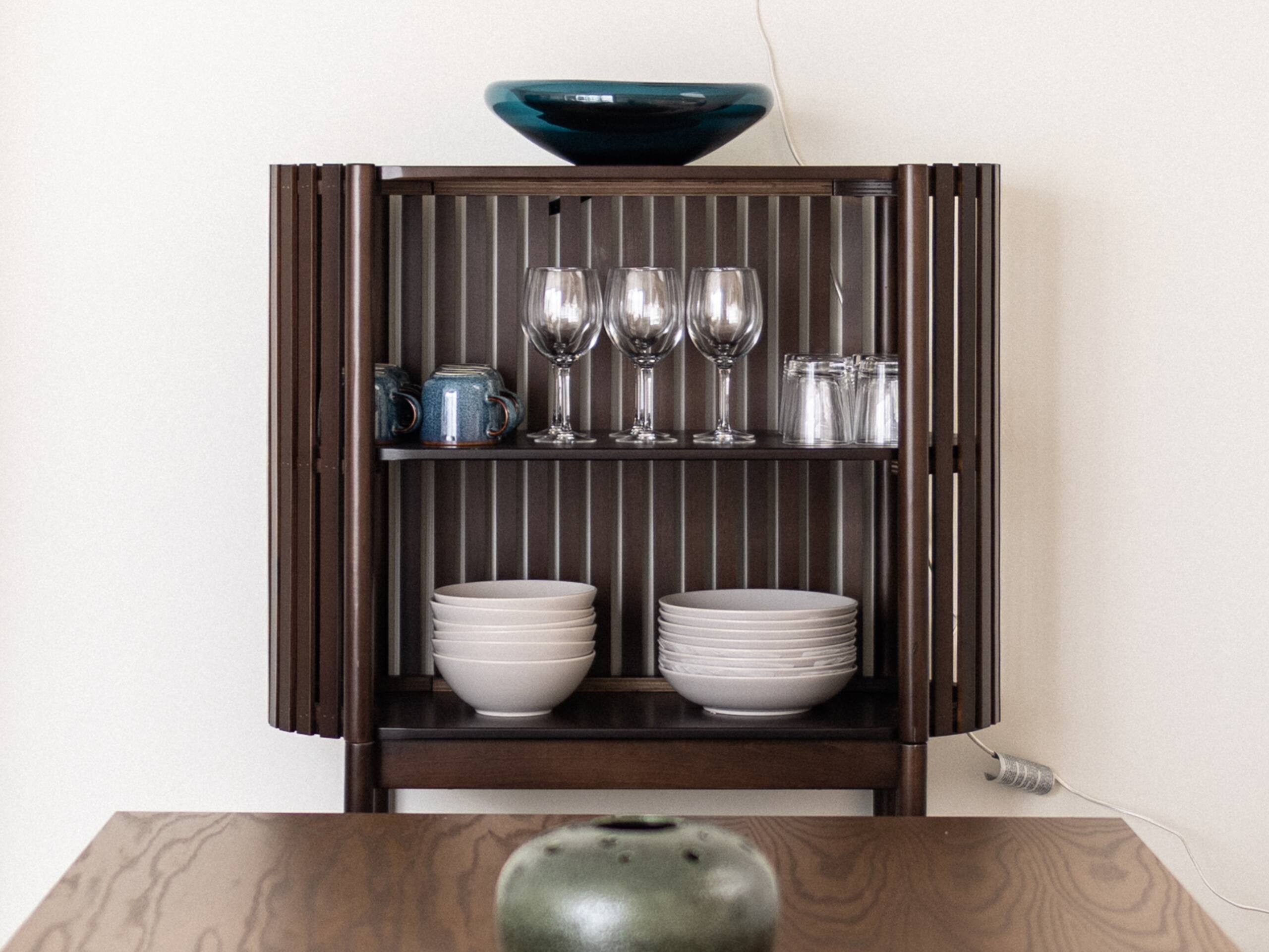 Close-up of the wooden cabinet with wine glasses, mugs, tumblers, bowls and plates beside the dining table